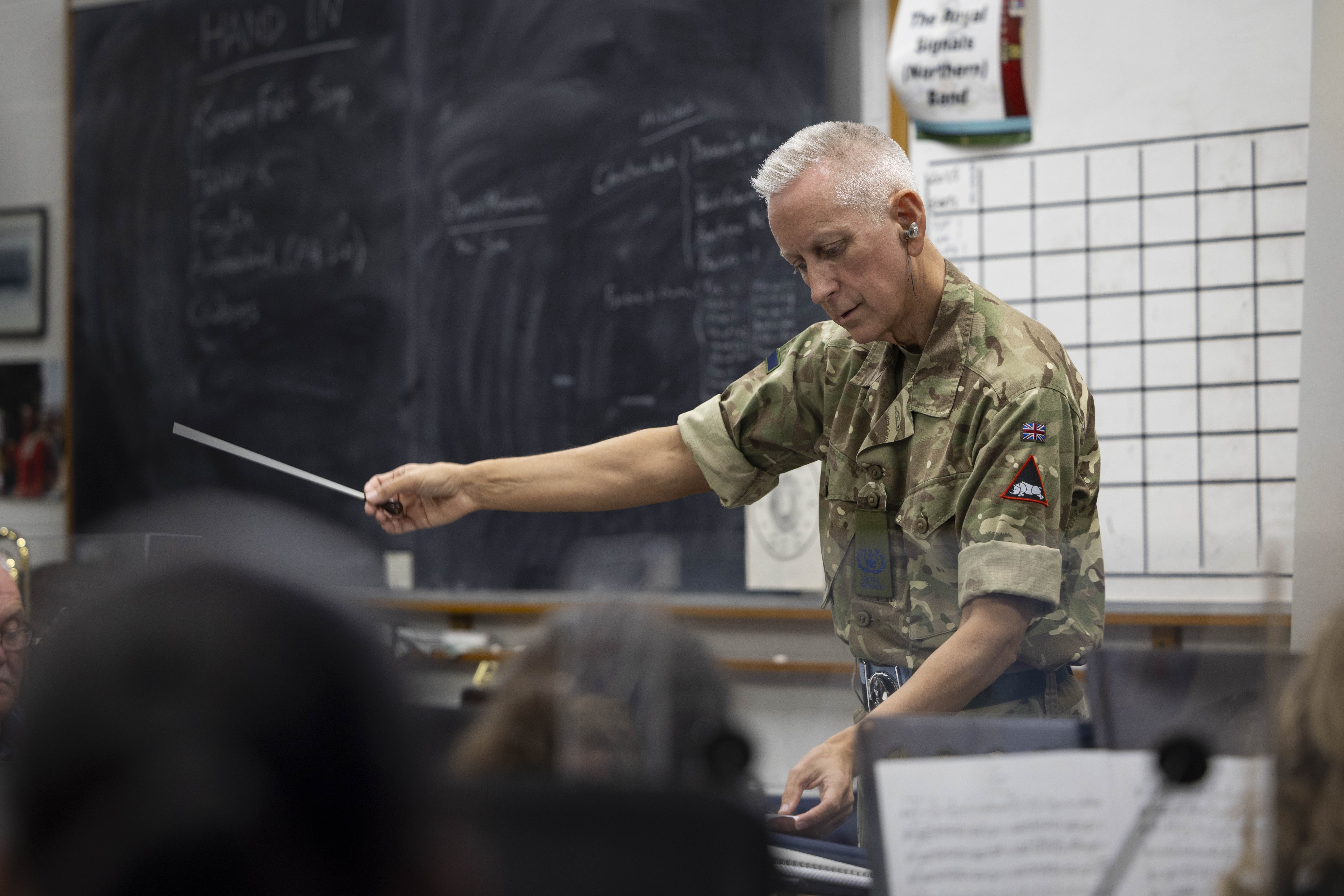 A person in a military camouflage uniform conducts a band with a baton in a classroom. A chalkboard and a chart are visible in the background. Musical instruments are seen in the foreground.