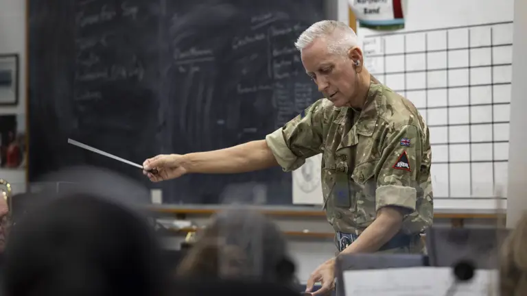 A person in a military camouflage uniform conducts a band with a baton in a classroom. A chalkboard and a chart are visible in the background. Musical instruments are seen in the foreground.