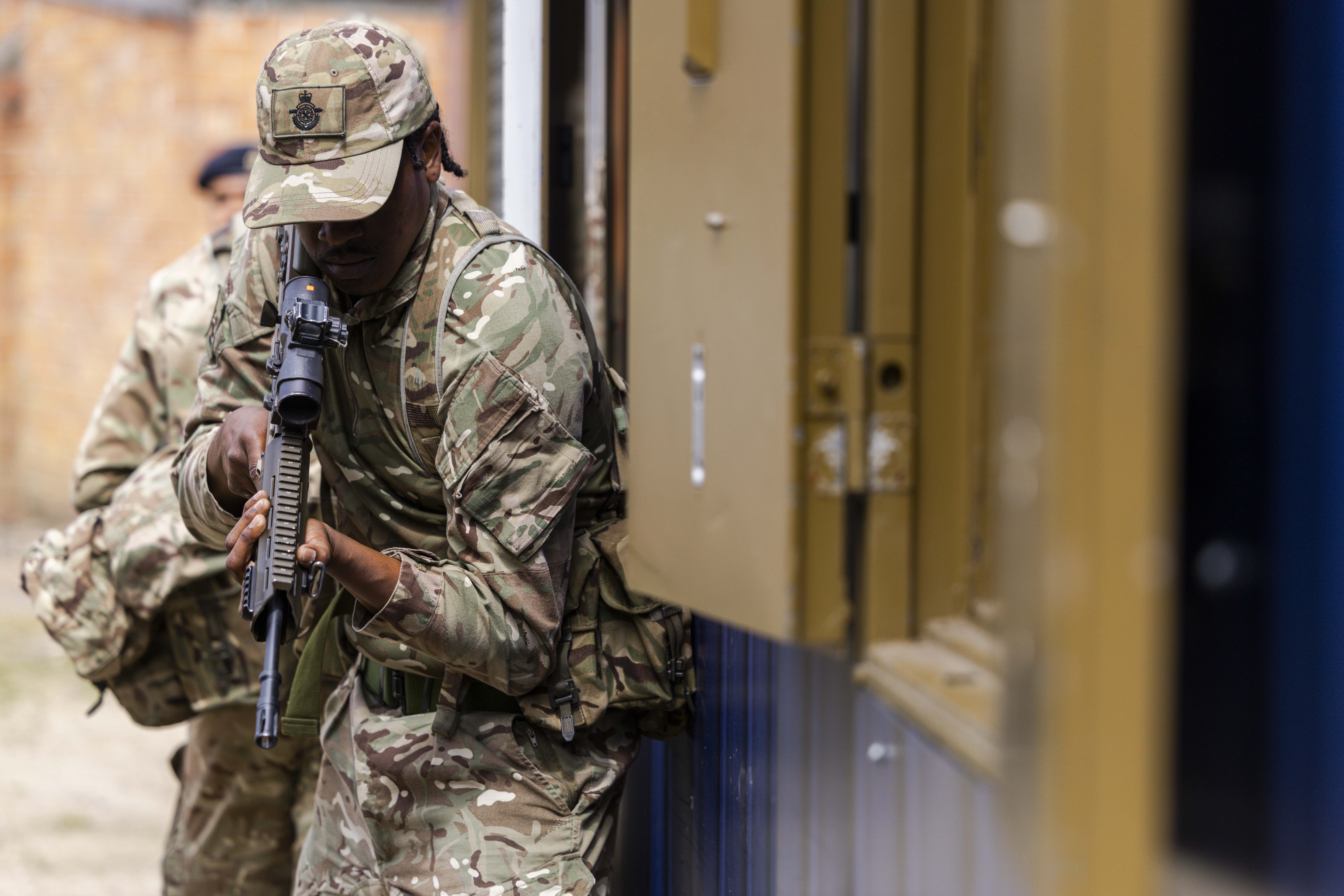 A person in camouflage military uniform holds a rifle while leaning against a building structure, ready for action.