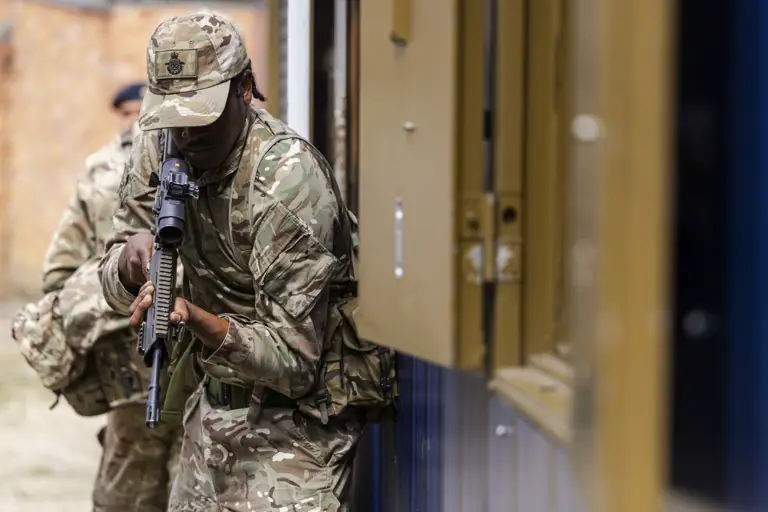 A person in camouflage military uniform holds a rifle while leaning against a building structure, ready for action.