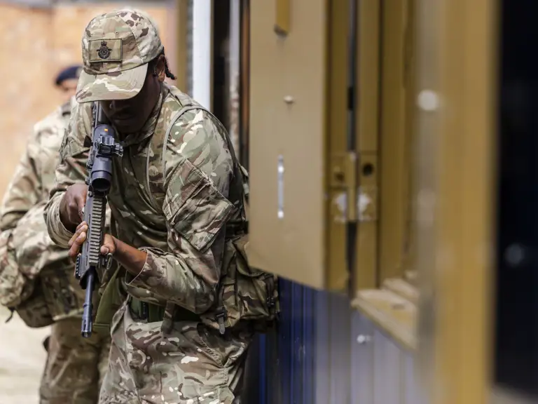 A person in camouflage military uniform holds a rifle while leaning against a building structure, ready for action.