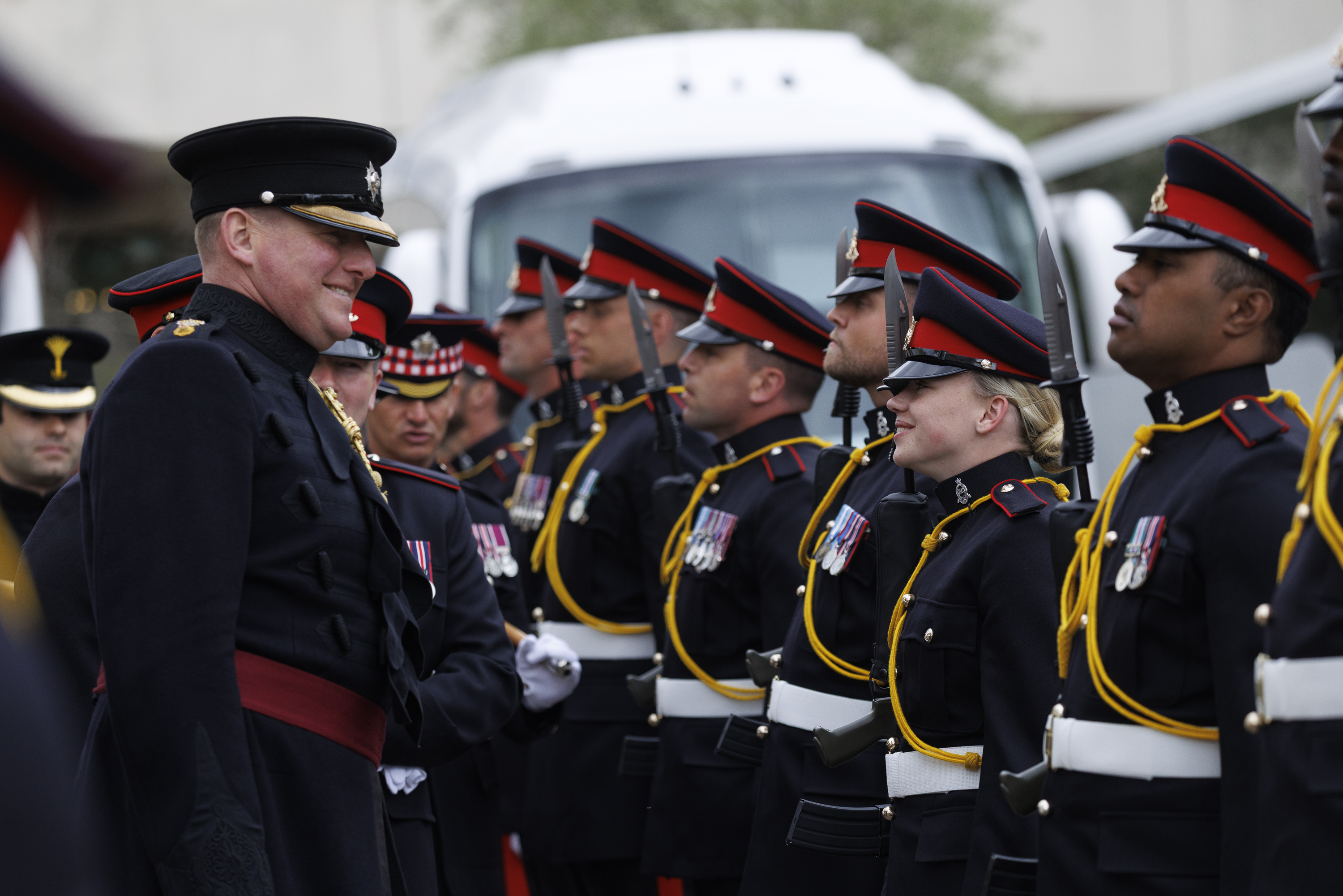 Military personnel in ceremonial dress uniforms, featuring black coats and red accents, are standing in formation.