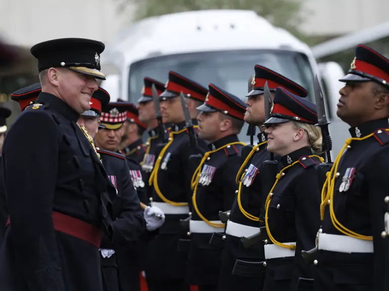 Military personnel in ceremonial dress uniforms, featuring black coats and red accents, are standing in formation.