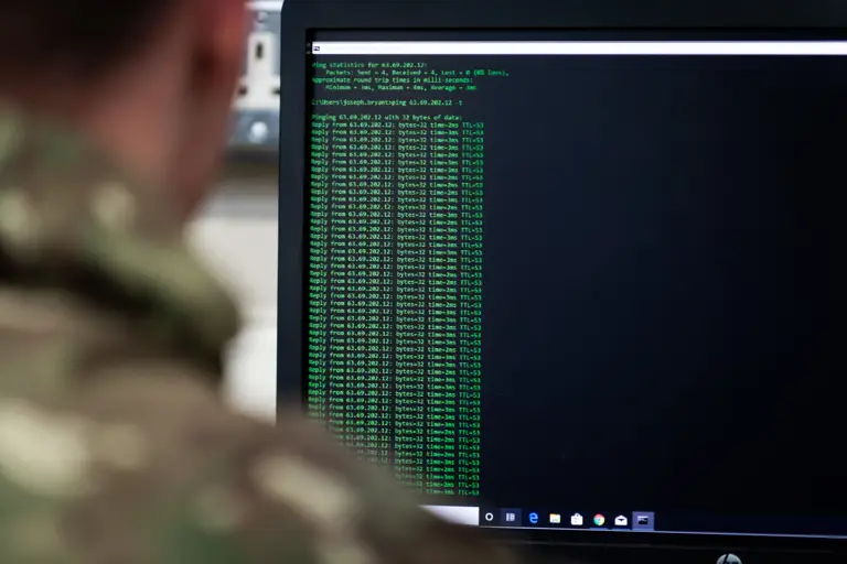 Person in camouflage uniform viewing a computer screen displaying network ping test results in green text.