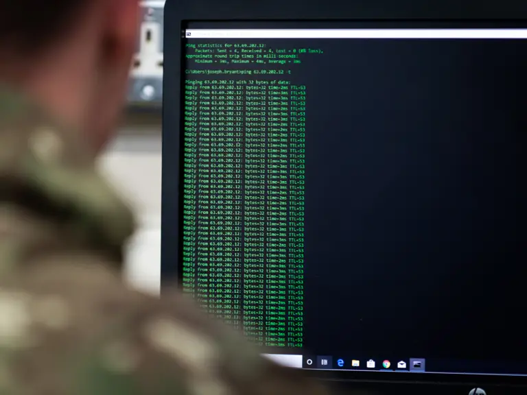 Person in camouflage uniform viewing a computer screen displaying network ping test results in green text.