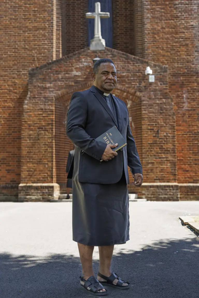 Clergyman dressed in a black suit jacket holding a Bible outside a brick church with a cross above the entrance.