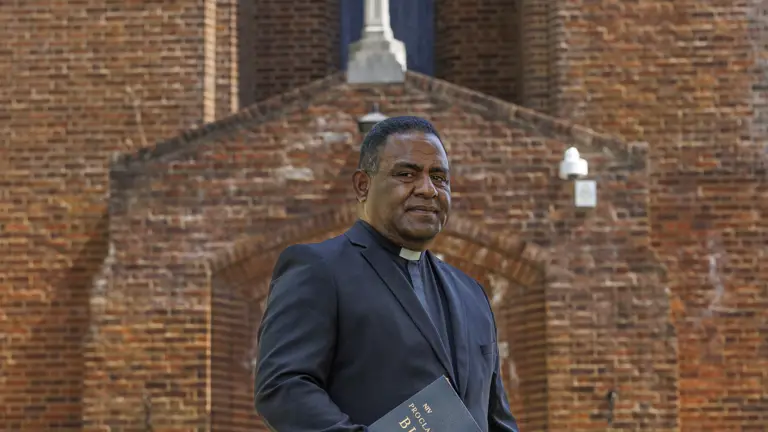Clergyman dressed in a black suit jacket holding a Bible outside a brick church with a cross above the entrance.