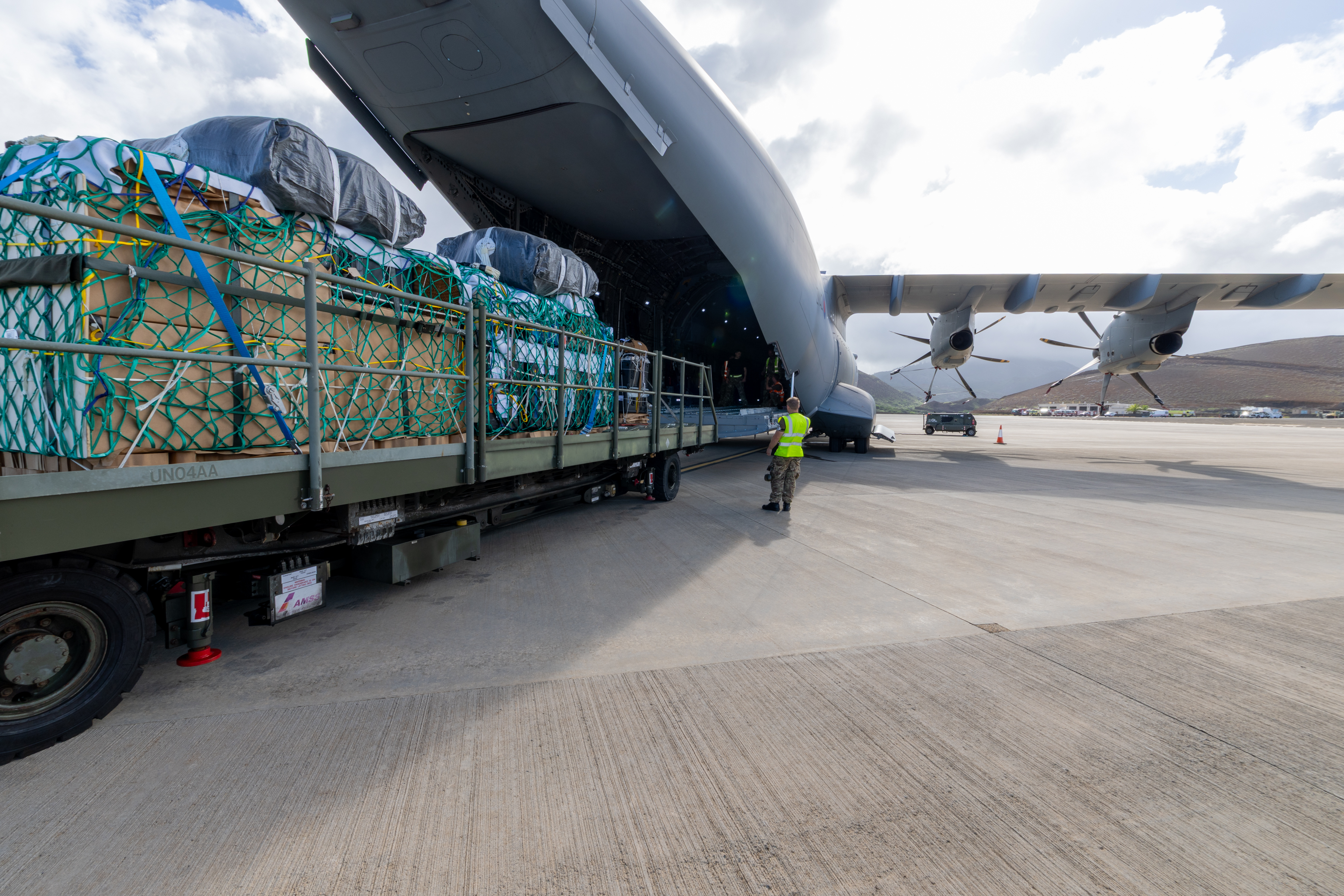 A large aircraft with its cargo door open is being loaded with supplies secured on a metal platform. A person in a high-visibility vest guides the loading process at an airport. The sky is partly cloudy.