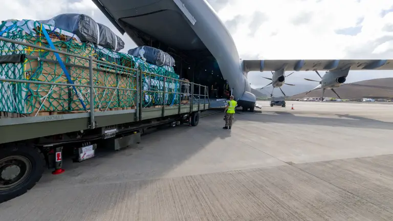 A large aircraft with its cargo door open is being loaded with supplies secured on a metal platform. A person in a high-visibility vest guides the loading process at an airport. The sky is partly cloudy.