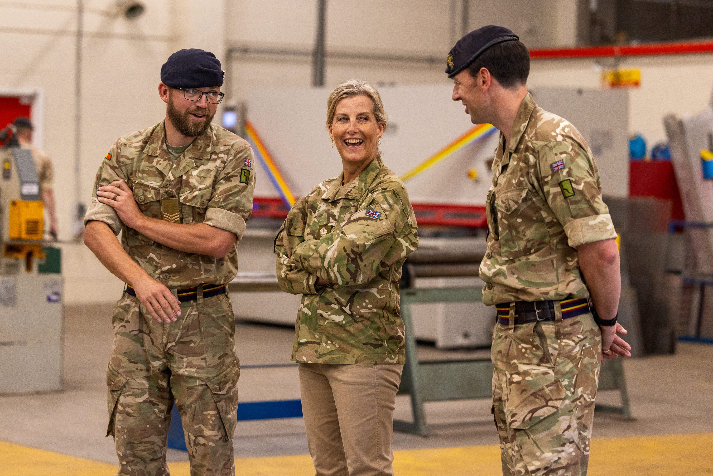 The Duchess of Edinburgh is seen wearing trousers and a camouflage print jacket stood inside with two soldiers in camouflage uniform either side of her.