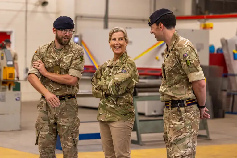 The Duchess of Edinburgh is seen wearing trousers and a camouflage print jacket stood inside with two soldiers in camouflage uniform either side of her.