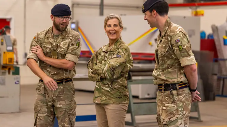 The Duchess of Edinburgh is seen wearing trousers and a camouflage print jacket stood inside with two soldiers in camouflage uniform either side of her.