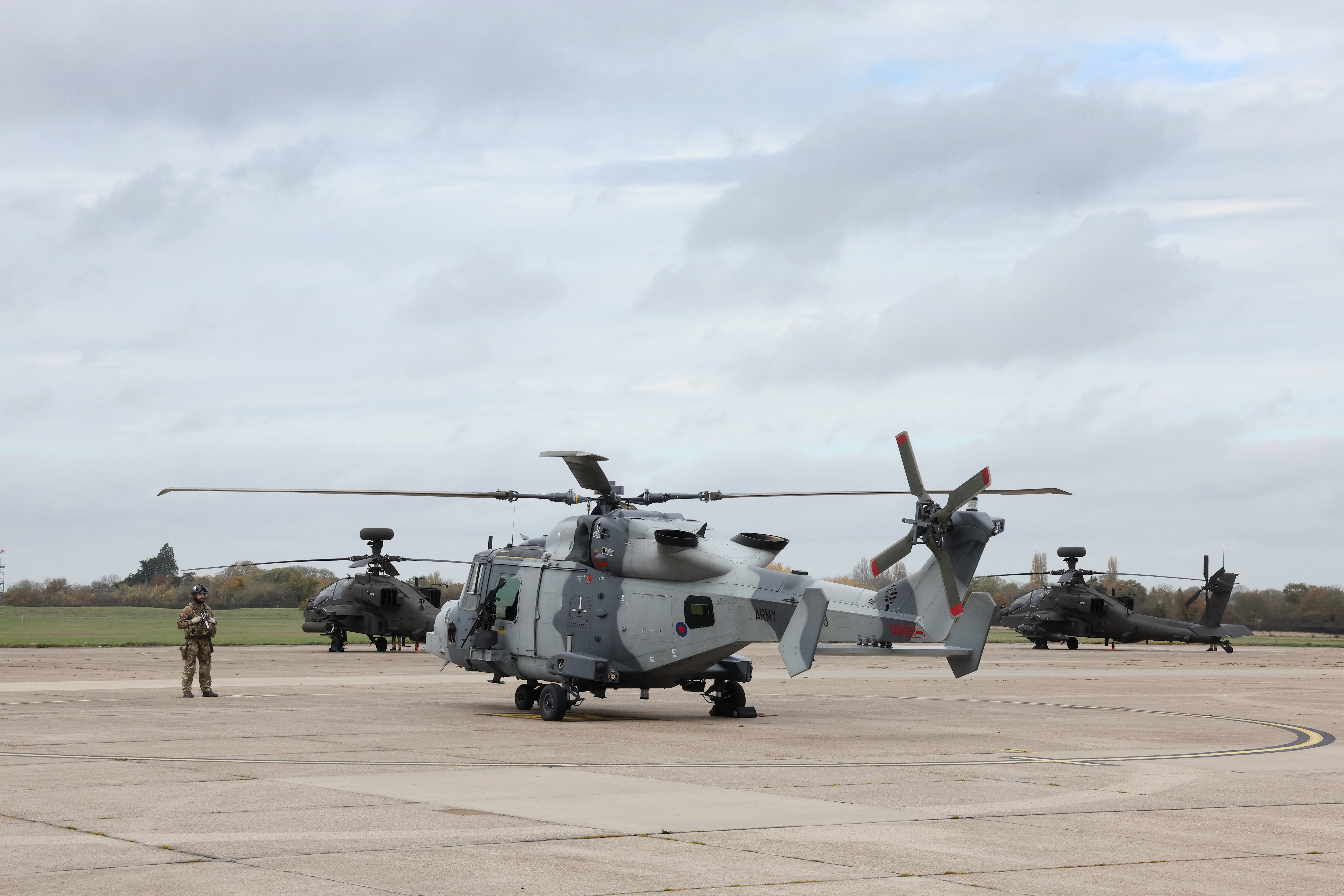 Military helicopters parked on a concrete airfield with a soldier standing nearby under a cloudy sky.