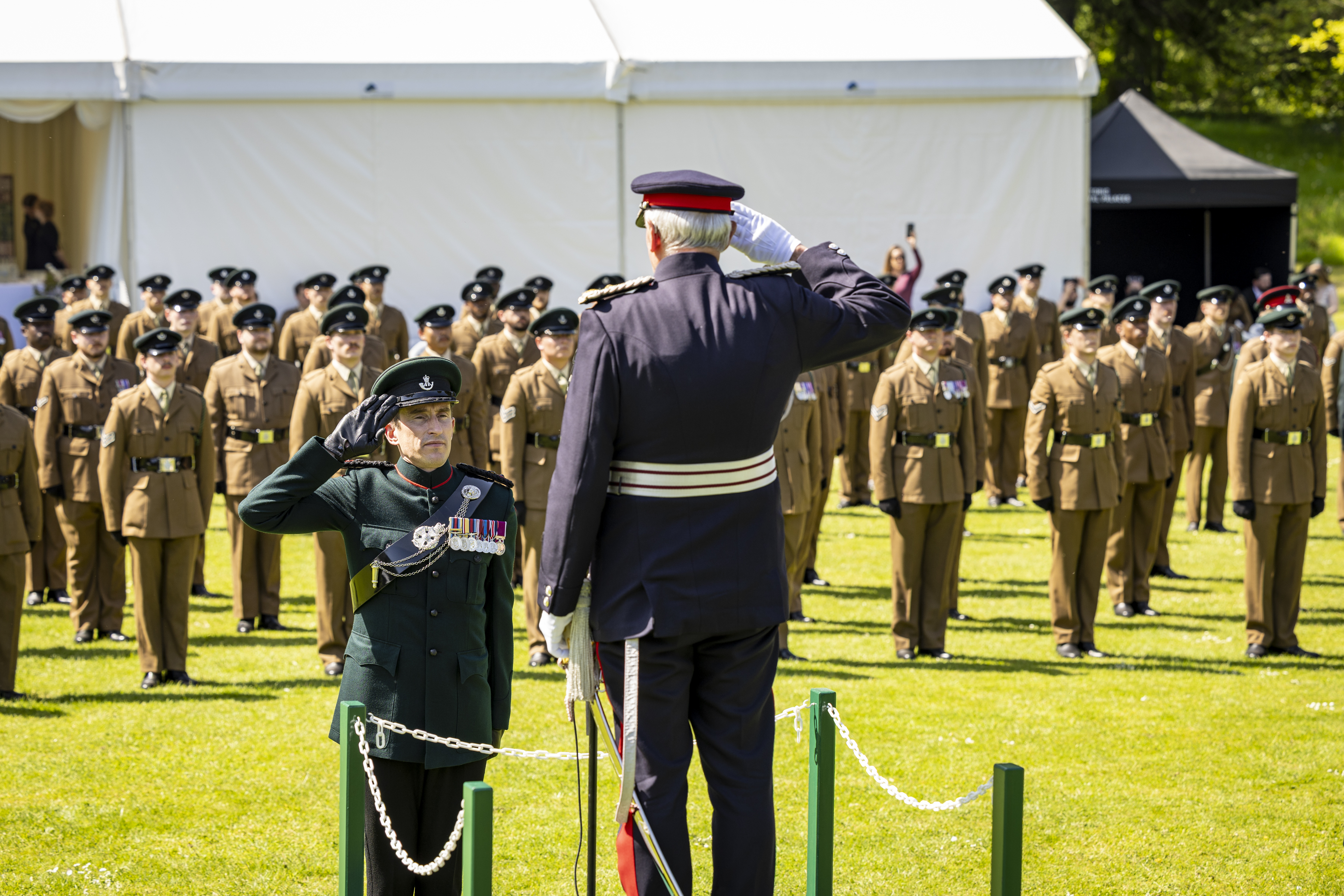 A formal military ceremony with uniformed personnel standing in formation on a lawn. Two individuals in the foreground exchange a salute, with one elevated on a small platform. A white tent and a green grassy area are visible in the background.