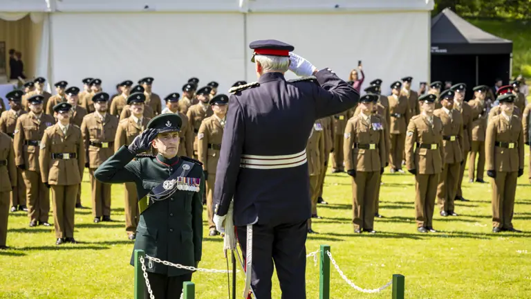 A formal military ceremony with uniformed personnel standing in formation on a lawn. Two individuals in the foreground exchange a salute, with one elevated on a small platform. A white tent and a green grassy area are visible in the background.