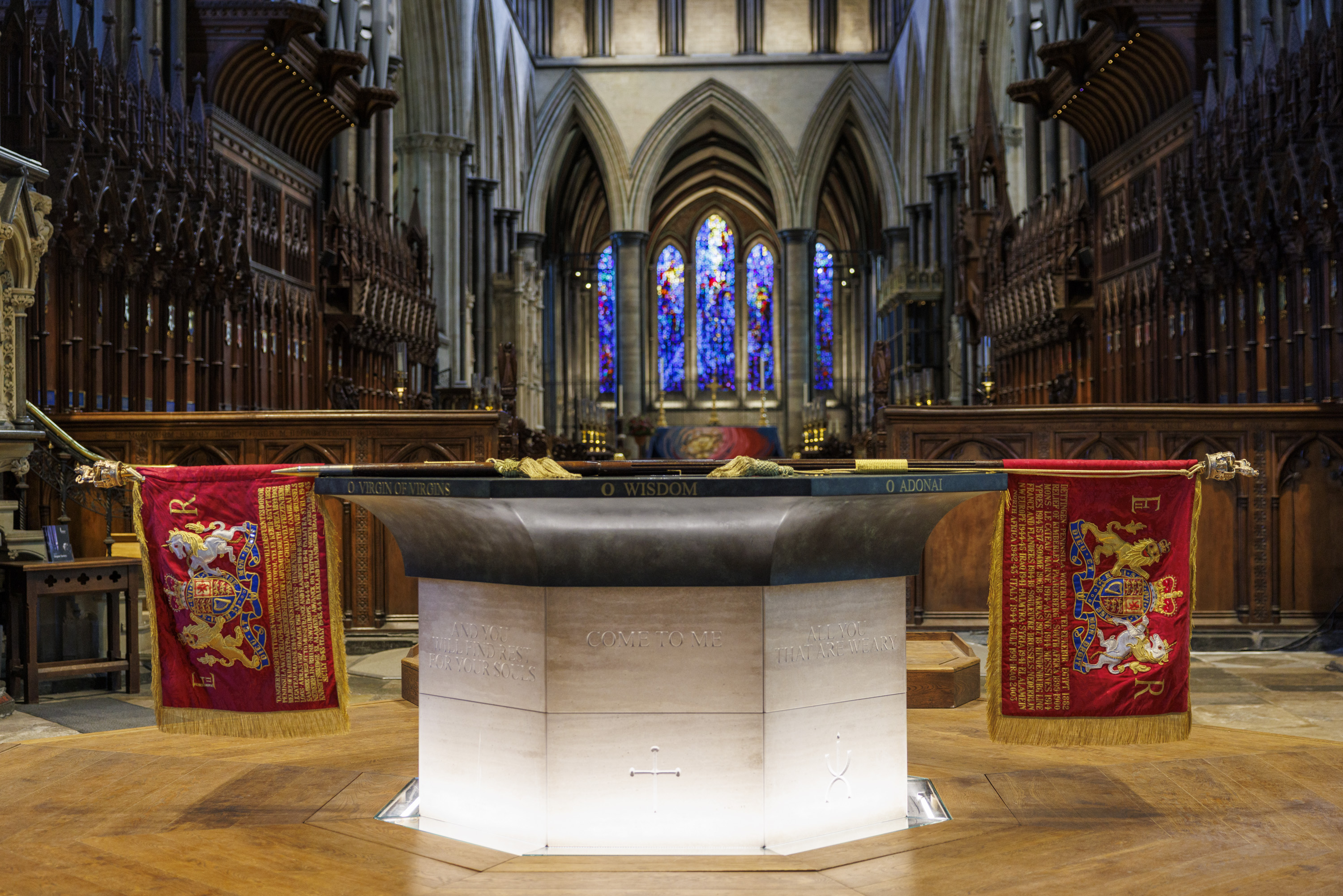 Stone baptismal font with inscriptions and red royal banners inside a cathedral featuring stained glass windows and wooden choir stalls.