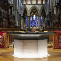 Stone baptismal font with inscriptions and red royal banners inside a cathedral featuring stained glass windows and wooden choir stalls.