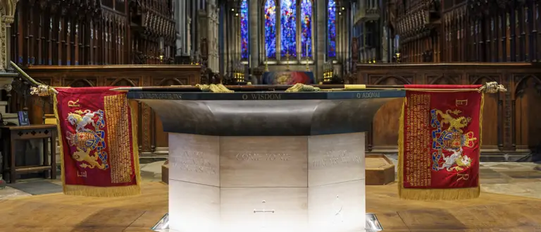Stone baptismal font with inscriptions and red royal banners inside a cathedral featuring stained glass windows and wooden choir stalls.