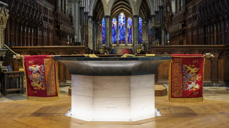 Stone baptismal font with inscriptions and red royal banners inside a cathedral featuring stained glass windows and wooden choir stalls.