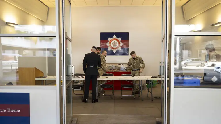 Four people in military uniforms stand around a table inside a room with a police emblem on the wall. The mood is focused and professional.