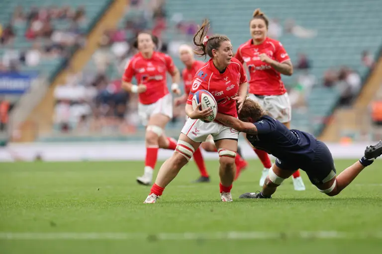 A women's rugby player in red dodges a tackle from a player in blue during a match. Teammates follow closely, showing focus and intensity.