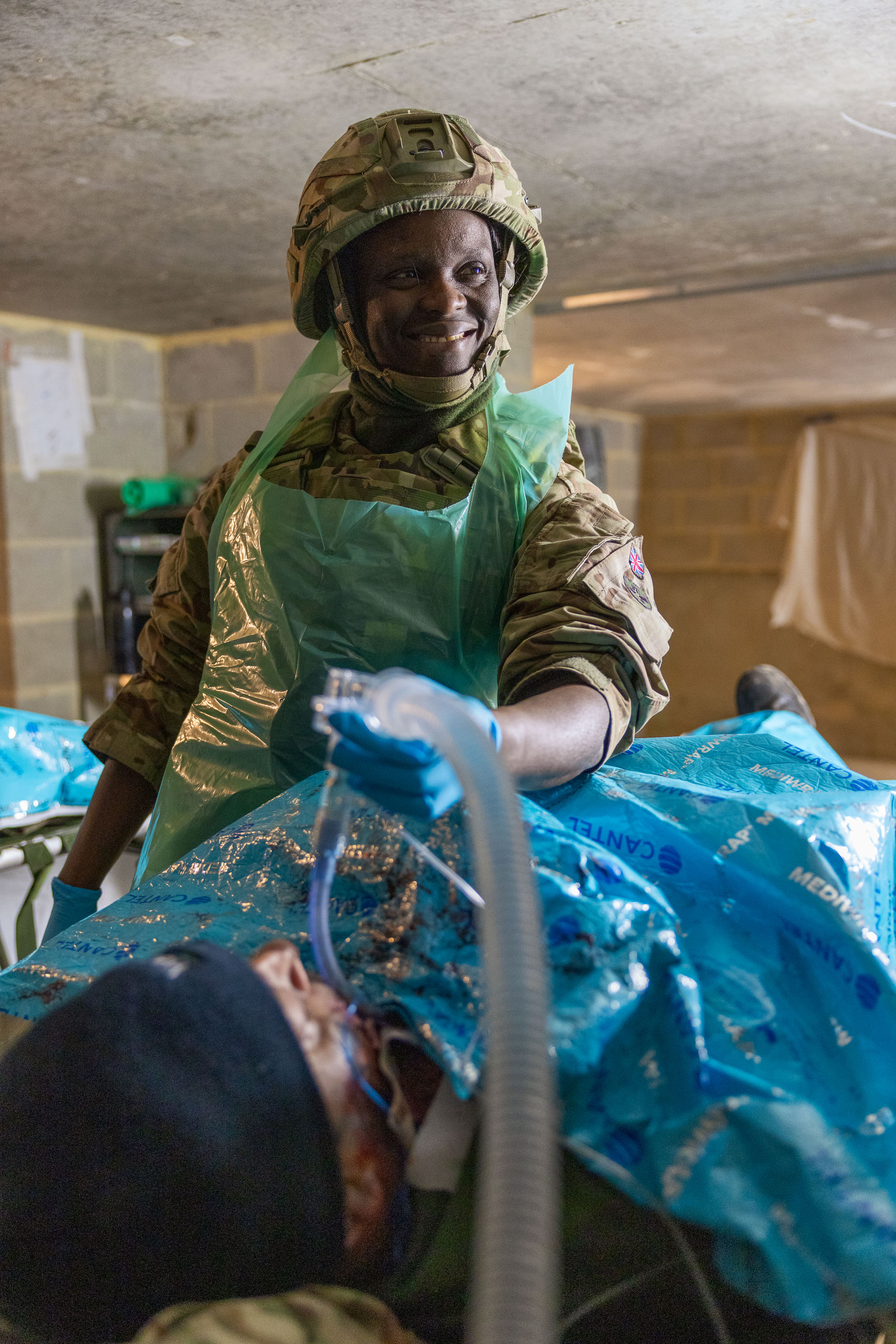 Military medic in camouflage uniform and protective apron attending to a patient covered with a blue medical sheet.