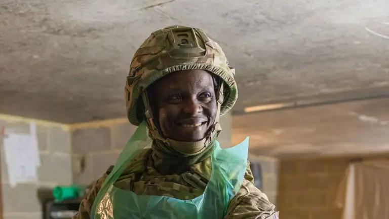 Military medic in camouflage uniform and protective apron attending to a patient covered with a blue medical sheet.