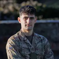 A person in a camouflage military uniform stands with arms crossed, looking forward. The background outside and blurred.