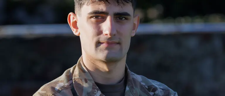 A person in a camouflage military uniform stands with arms crossed, looking forward. The background outside and blurred.