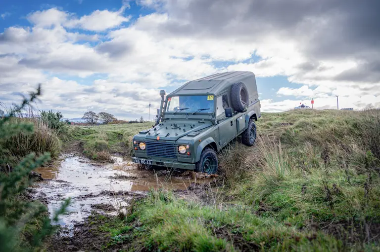 A green off-road vehicle navigates a muddy puddle on a grassy terrain under a partially cloudy sky. The scene conveys adventure and rugged exploration.