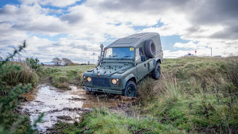 A green off-road vehicle navigates a muddy puddle on a grassy terrain under a partially cloudy sky. The scene conveys adventure and rugged exploration.