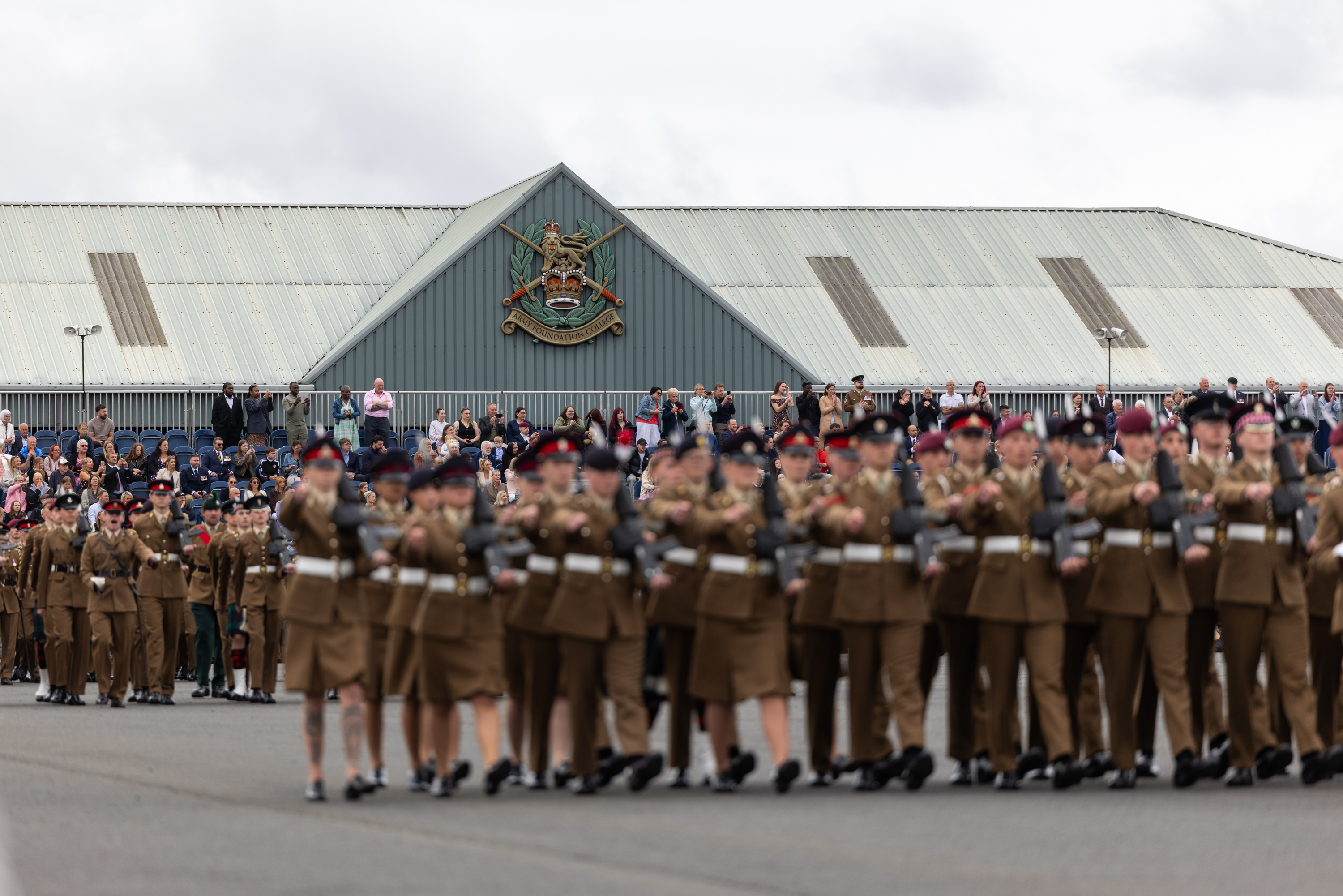 A large group of uniformed individuals are marching in formation on a paved surface. Spectators are gathered to watch the procession. A building in the background features a prominent emblem and is topped with a corrugated metal roof.