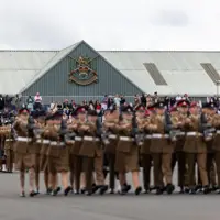 A large group of uniformed individuals are marching in formation on a paved surface. Spectators are gathered to watch the procession. A building in the background features a prominent emblem and is topped with a corrugated metal roof.