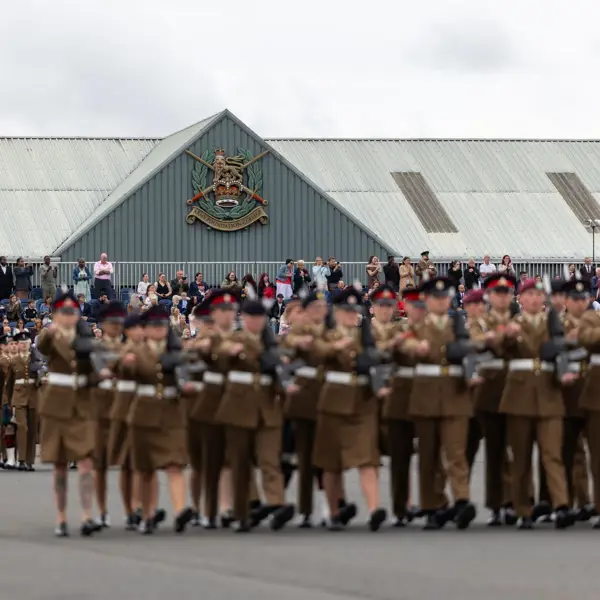 A large group of uniformed individuals are marching in formation on a paved surface. Spectators are gathered to watch the procession. A building in the background features a prominent emblem and is topped with a corrugated metal roof.