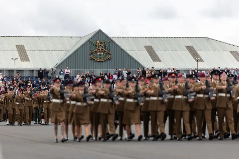 A large group of uniformed individuals are marching in formation on a paved surface. Spectators are gathered to watch the procession. A building in the background features a prominent emblem and is topped with a corrugated metal roof.