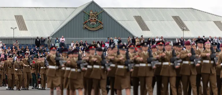 A large group of uniformed individuals are marching in formation on a paved surface. Spectators are gathered to watch the procession. A building in the background features a prominent emblem and is topped with a corrugated metal roof.