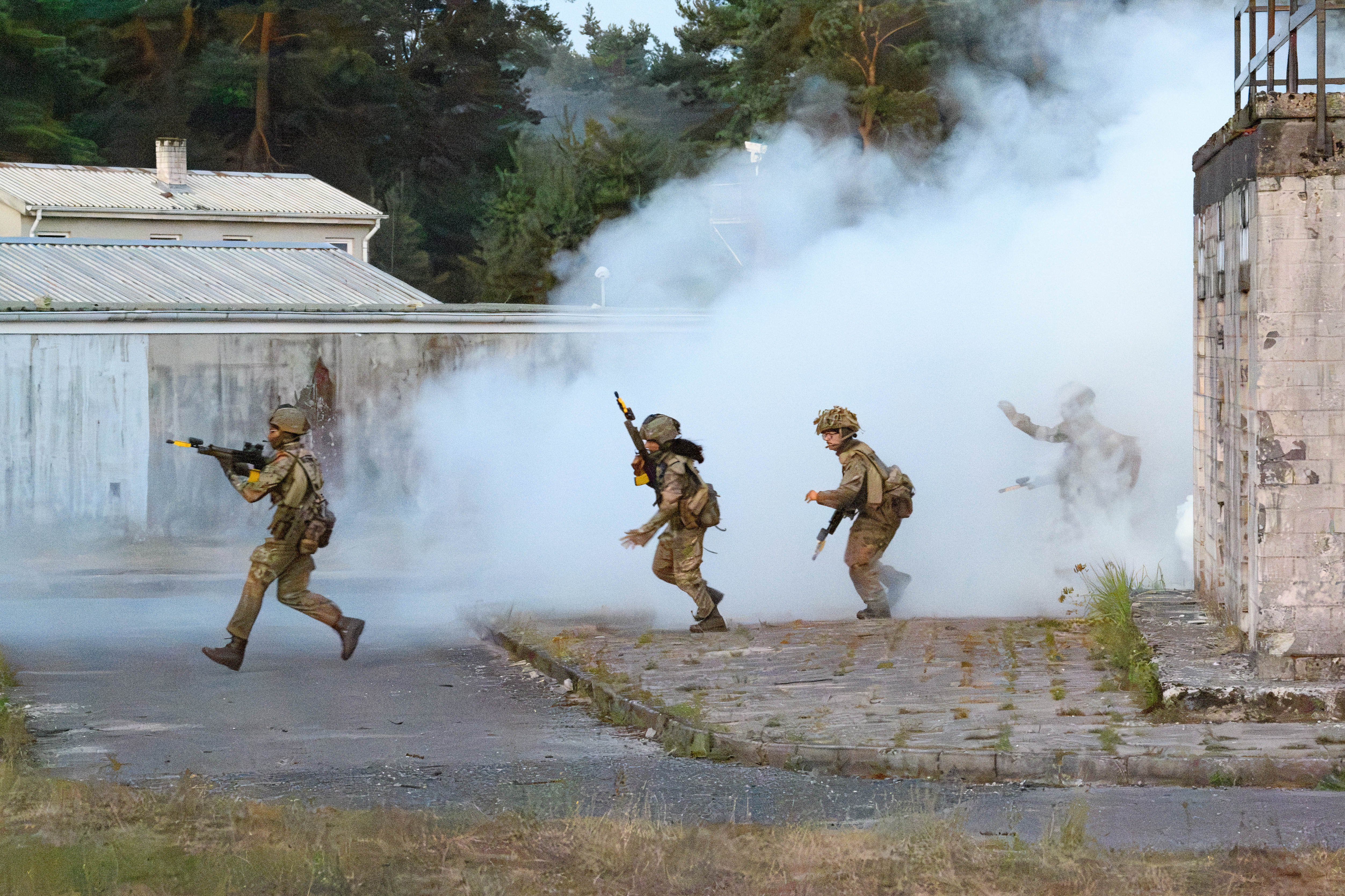 A group of soldiers in tactical gear move through a training ground with thick smoke surrounding the area. They carry rifles and appear to be in the midst of a training exercise near a wooded area and industrial-style buildings.