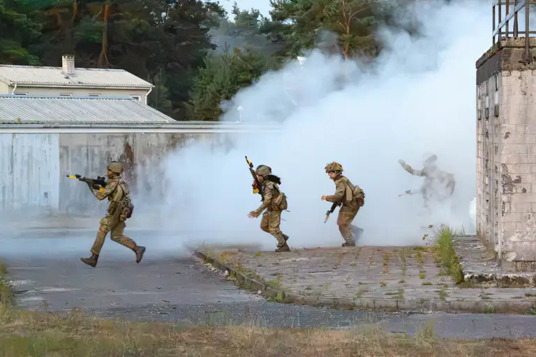 A group of soldiers in tactical gear move through a training ground with thick smoke surrounding the area. They carry rifles and appear to be in the midst of a training exercise near a wooded area and industrial-style buildings.