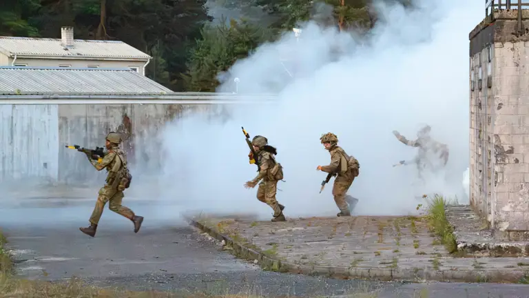 A group of soldiers in tactical gear move through a training ground with thick smoke surrounding the area. They carry rifles and appear to be in the midst of a training exercise near a wooded area and industrial-style buildings.