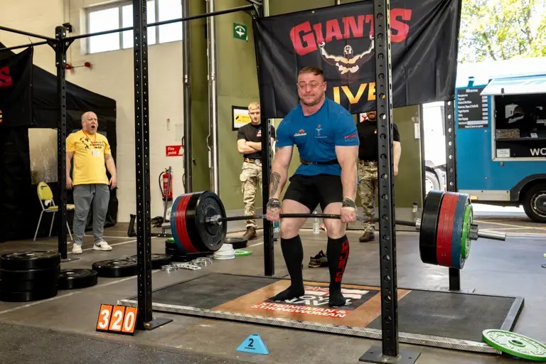 A man in a blue shirt deadlifts heavy weights in a gym, concentrating intensely. Spectators watch beside a 