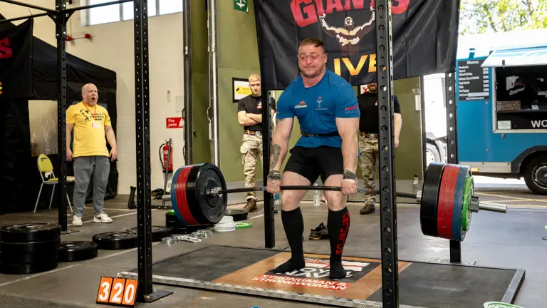 A man in a blue shirt deadlifts heavy weights in a gym, concentrating intensely. Spectators watch beside a