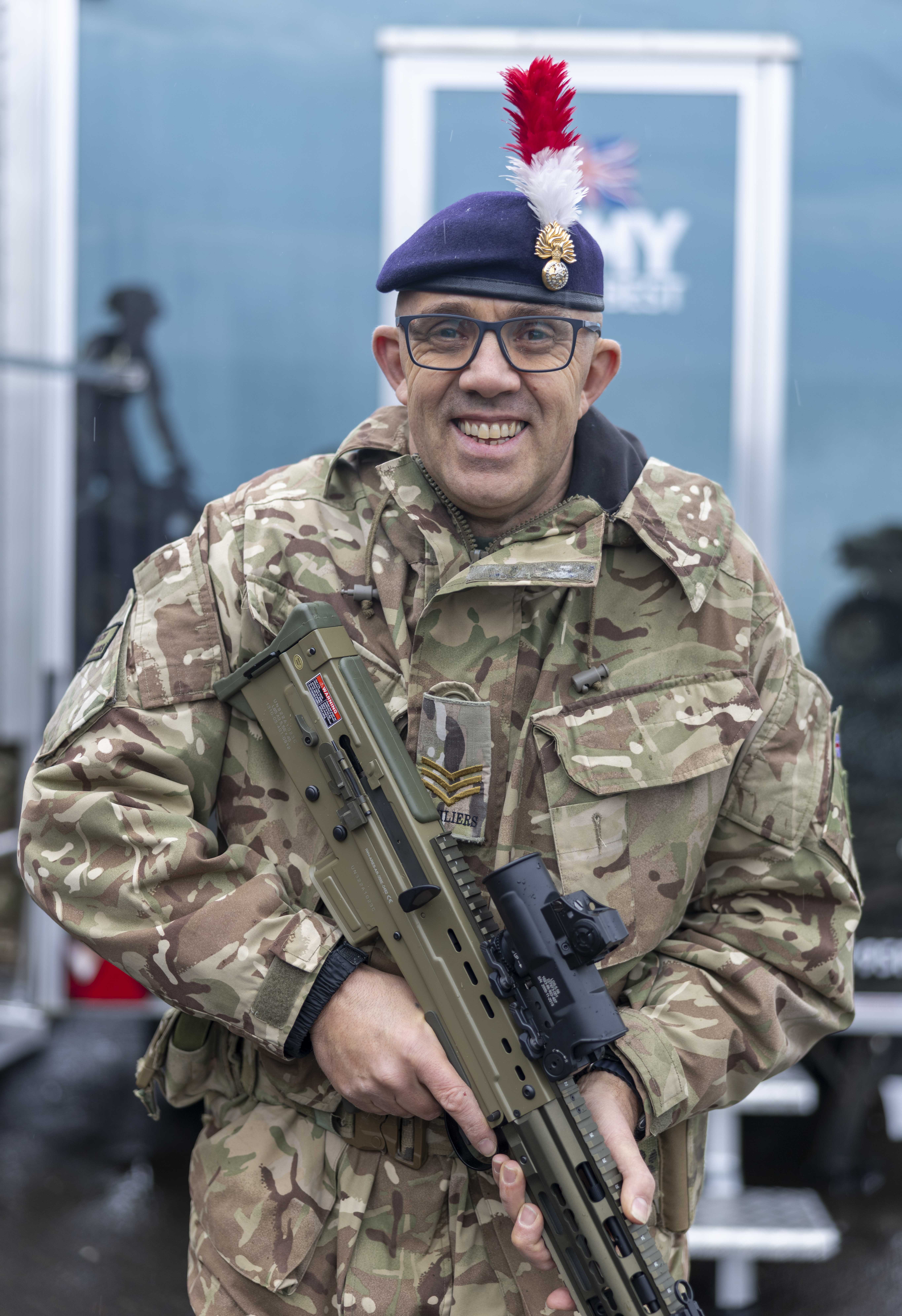 Soldier in camouflage uniform holding a modern assault rifle with a red and white plume on his beret.