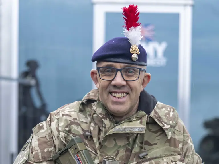 Soldier in camouflage uniform holding a modern assault rifle with a red and white plume on his beret.