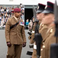 A person in military uniform with medals inspects a row of soldiers in formation. In the background, people are seated in bleachers watching the event.