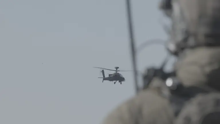 Image shows a AH-64E Apache helicopter mid flight in the sky. The foreground shows a soldier looking towards the helicopter with the perspective of the camera over his shoulder.
