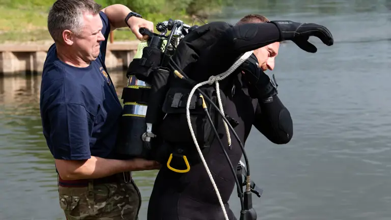 A man in camouflage trousers assists a diver wearing a black wetsuit and diving gear near a calm body of water.
