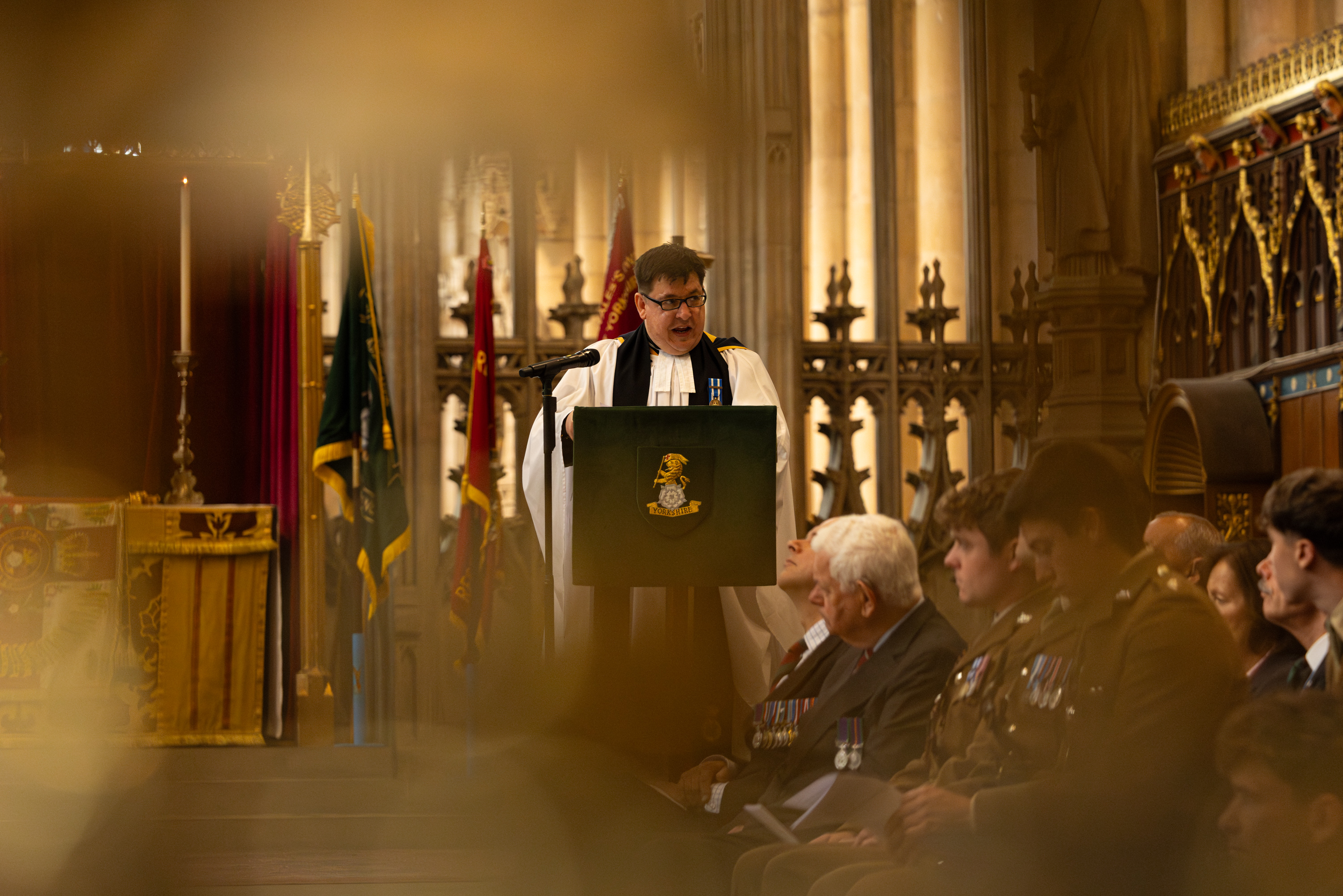 A religious ceremony taking place inside a grand church, featuring a clergy member speaking from a pulpit in front of a large altar. Attendees, including several in military uniforms, are seated and standing nearby. The background is adorned with ornate stained glass windows and religious symbols.