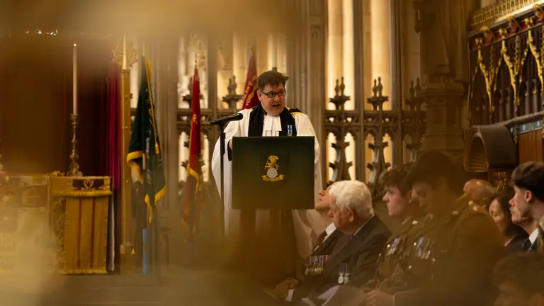 A religious ceremony taking place inside a grand church, featuring a clergy member speaking from a pulpit in front of a large altar. Attendees, including several in military uniforms, are seated and standing nearby. The background is adorned with ornate stained glass windows and religious symbols.