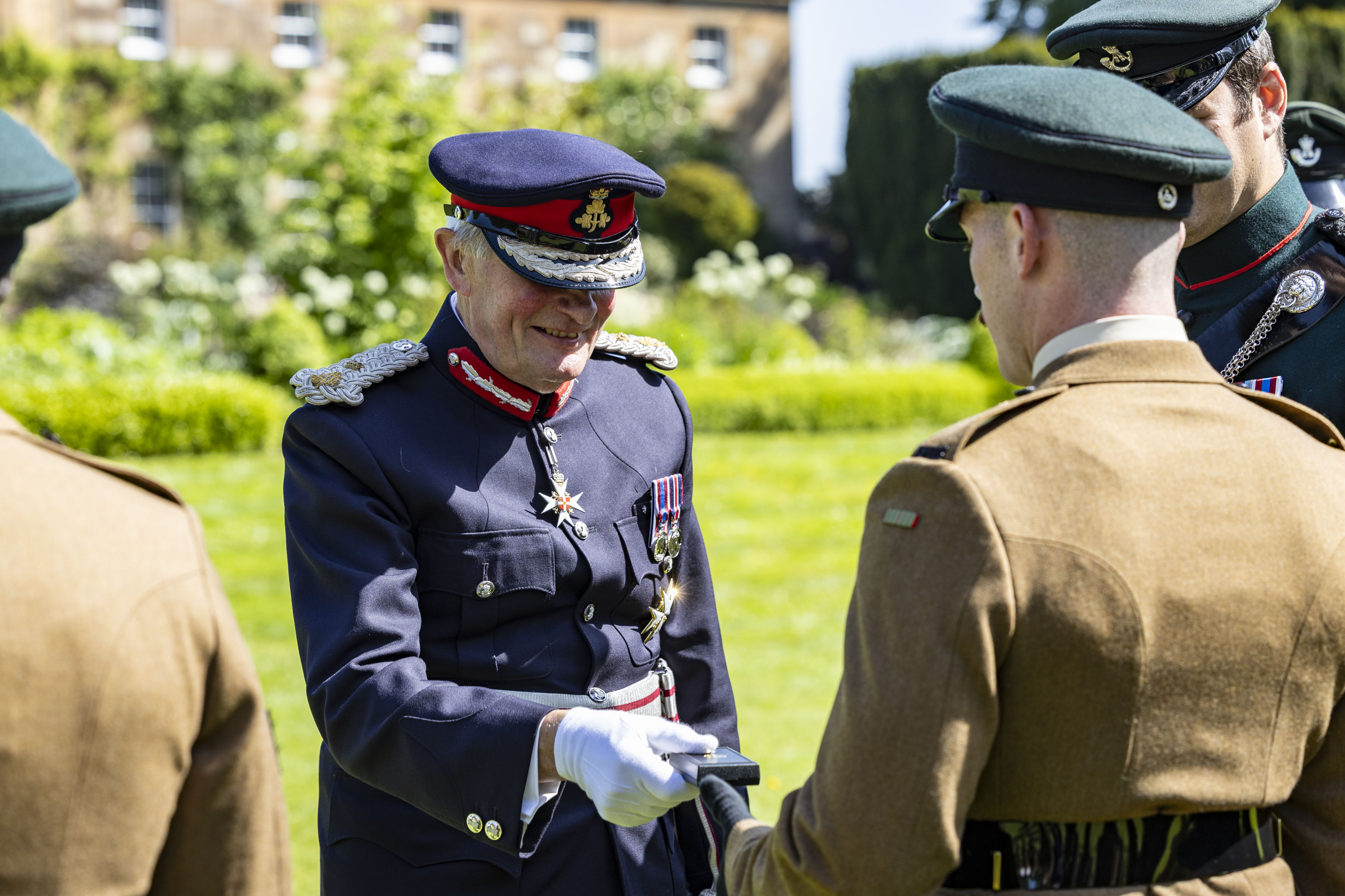 A person in a formal military uniform, adorned with medals, is shaking hands with another individual in military attire. Two additional individuals in similar uniforms are partially visible. They are outdoors, standing on a lawn with a building and greenery in the background.