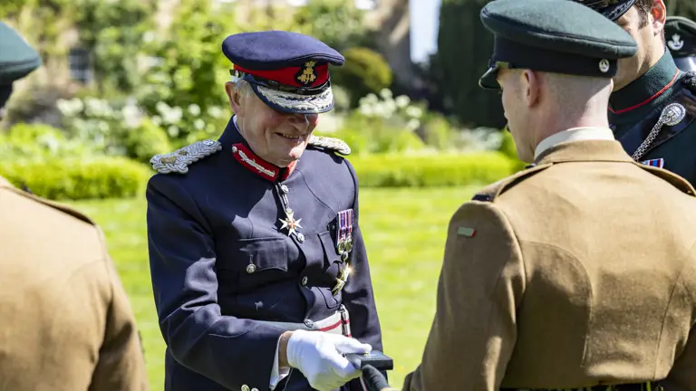 A person in a formal military uniform, adorned with medals, is shaking hands with another individual in military attire. Two additional individuals in similar uniforms are partially visible. They are outdoors, standing on a lawn with a building and greenery in the background.